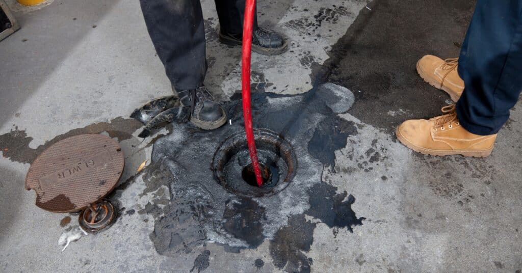 Plumber using a hydro jetting machine to clear a clogged drain in a commercial kitchen, showcasing grease buildup and maintenance of plumbing systems.