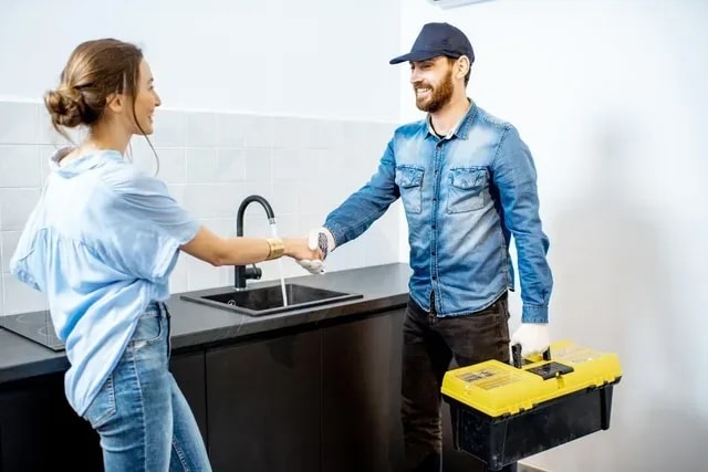 Plumber in uniform shaking hands with a homeowner in a modern kitchen, holding a toolbox, representing Plumbing Outfitters' professional service and customer engagement in North Austin.
