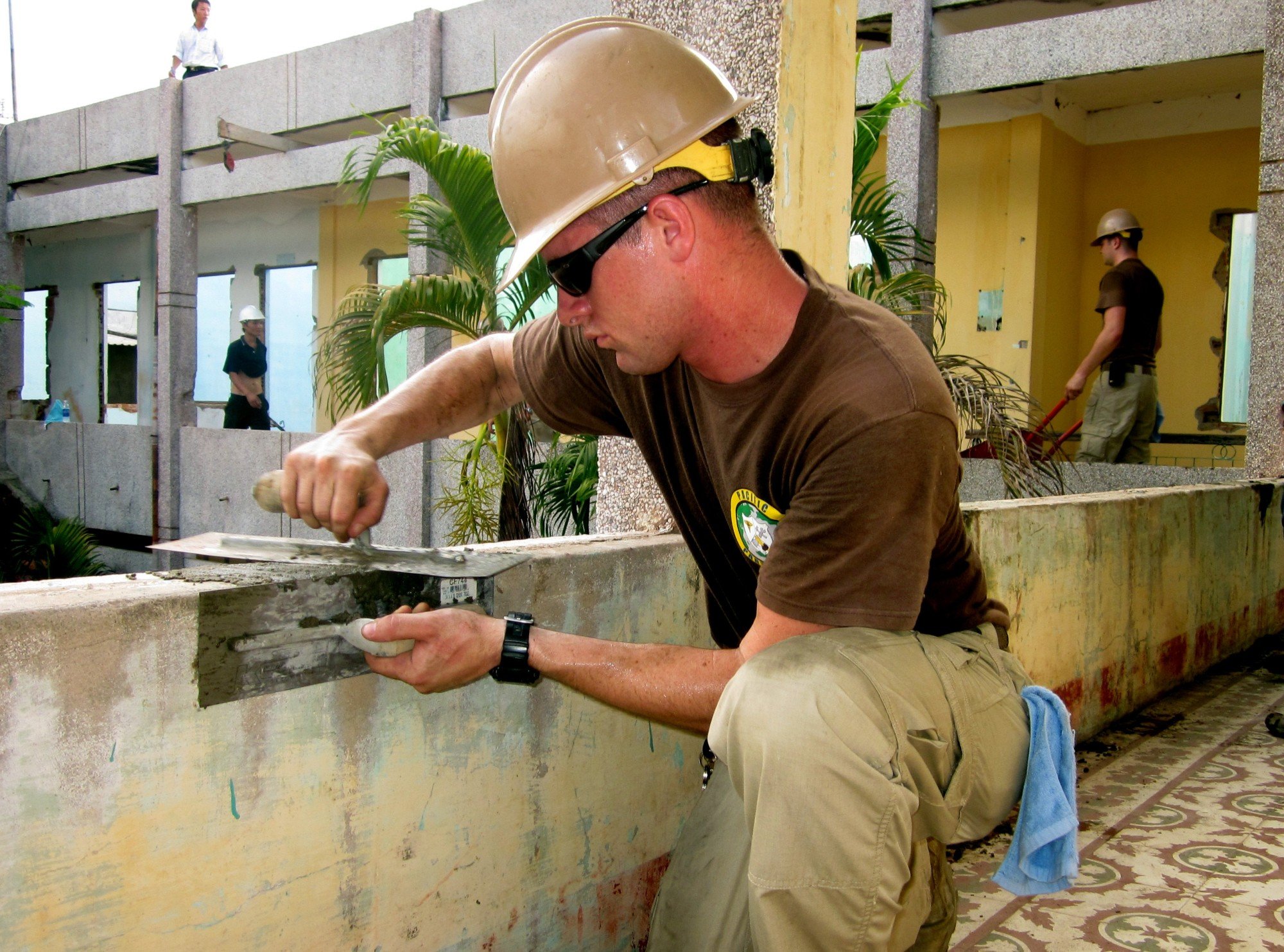 Construction worker applying cement to a wall, wearing a hard hat, with additional workers and greenery in the background, illustrating skilled labor in plumbing and construction services.