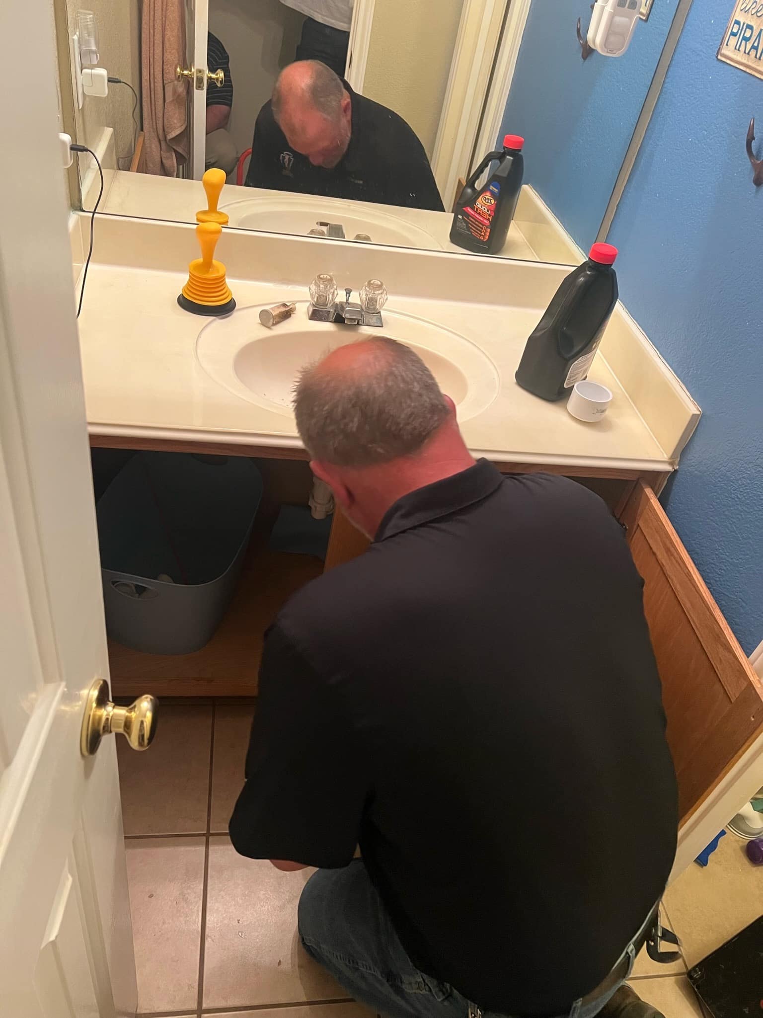 Plumber inspecting under bathroom sink, demonstrating expert plumbing service in residential setting, with tools and cleaning supplies visible.