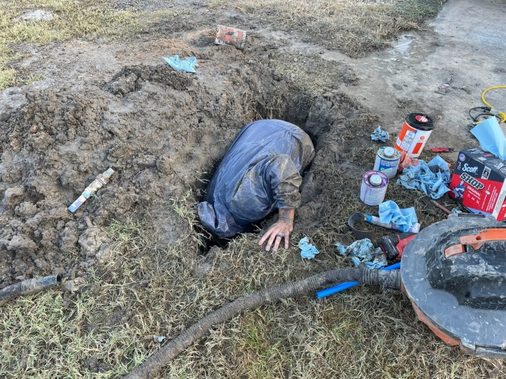 Plumber working in a muddy trench, repairing a water softener system, surrounded by tools and materials.
