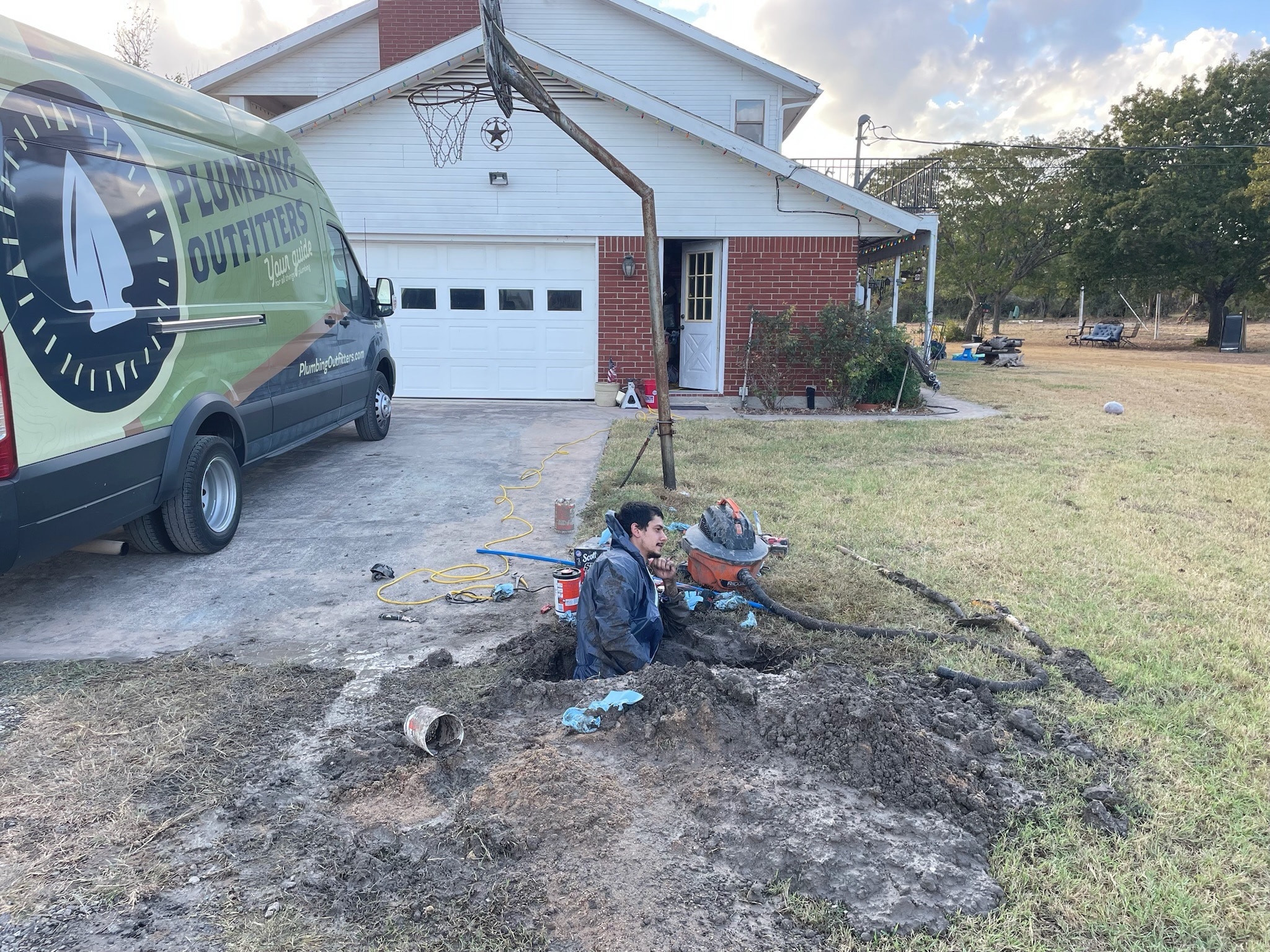 Plumbing Outfitters technician working in a trench near a home, with service van visible, addressing plumbing issues in Liberty Hill, TX.