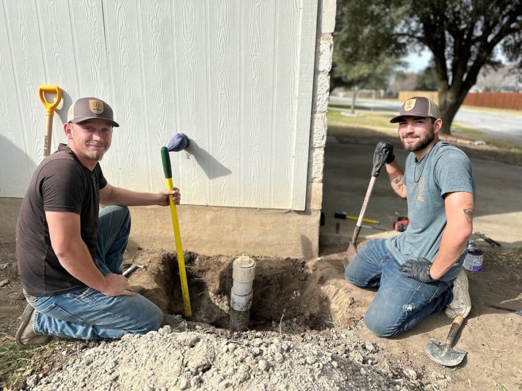 Two licensed plumbers working on a plumbing repair project in Cedar Park, using tools near a dug-out area around a pipe, showcasing hands-on expertise in addressing plumbing issues.