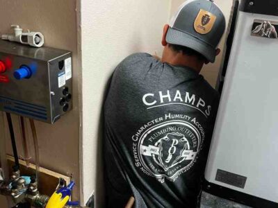 Plumber in gray shirt with "CHAMPS" text and Plumbing Outfitters logo working on plumbing system in residential setting, highlighting seamless repiping solutions in Taylor, TX.