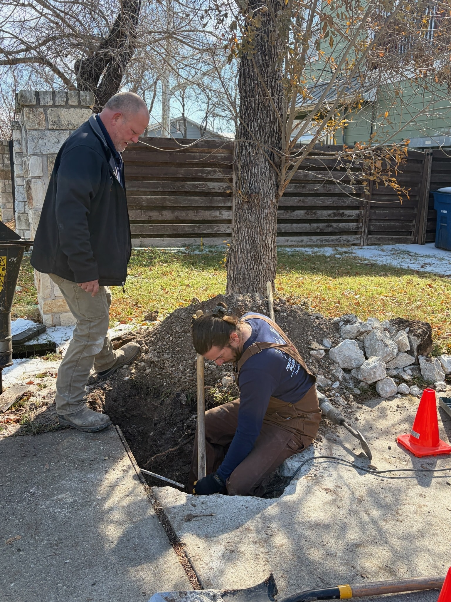 Two plumbers performing trenchless water line replacement, one digging in a hole while the other supervises, surrounded by tools and safety cones, showcasing modern plumbing techniques.